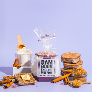 Packaged Dam Good English Muffins with cinnamon swirl, a mug of coffee, and cinnamon sticks on a purple background.